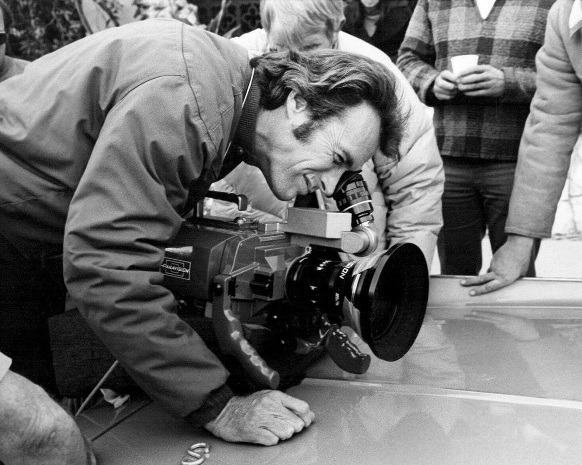 Clint Eastwood leaning over a car roof to look through a film camera, people watching behind him