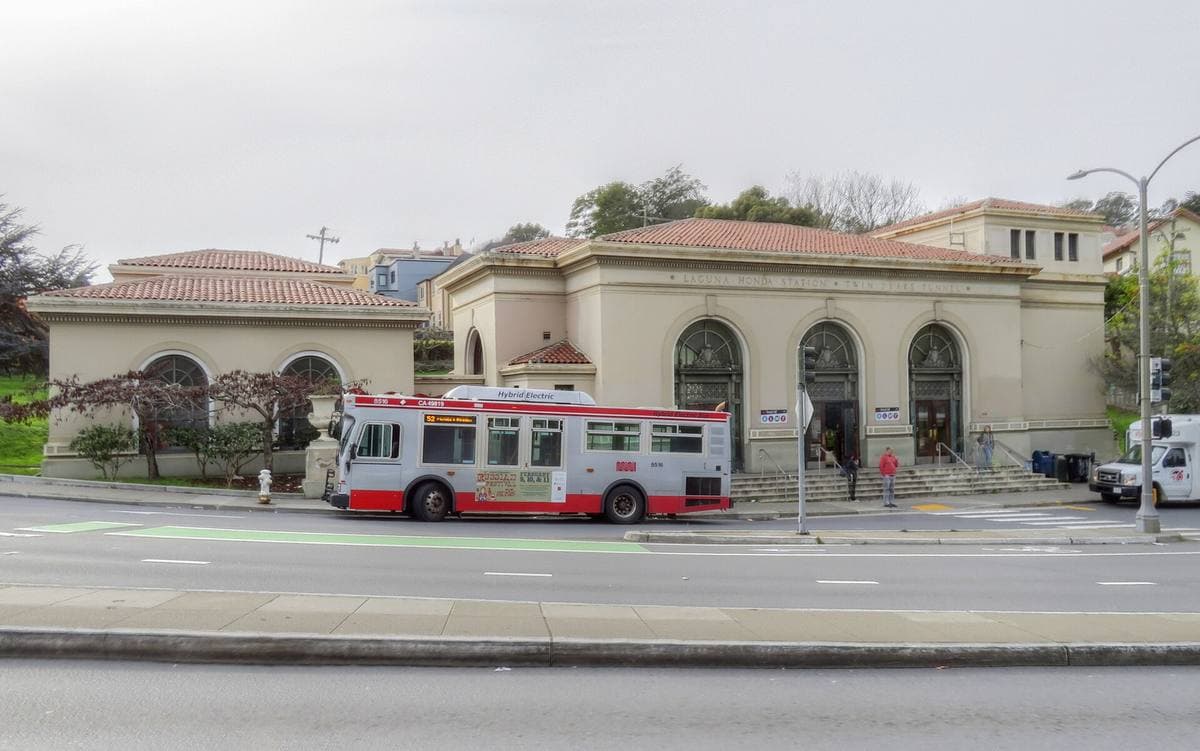 A low Spanish-style station building with arched windows and a red-and-white bus in front