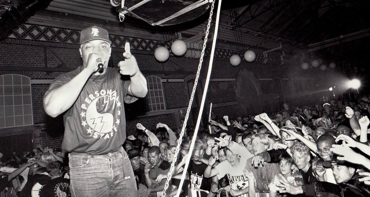 Chuck D in a cap with a microphone on stage above a crowd with raised hands, in black and white