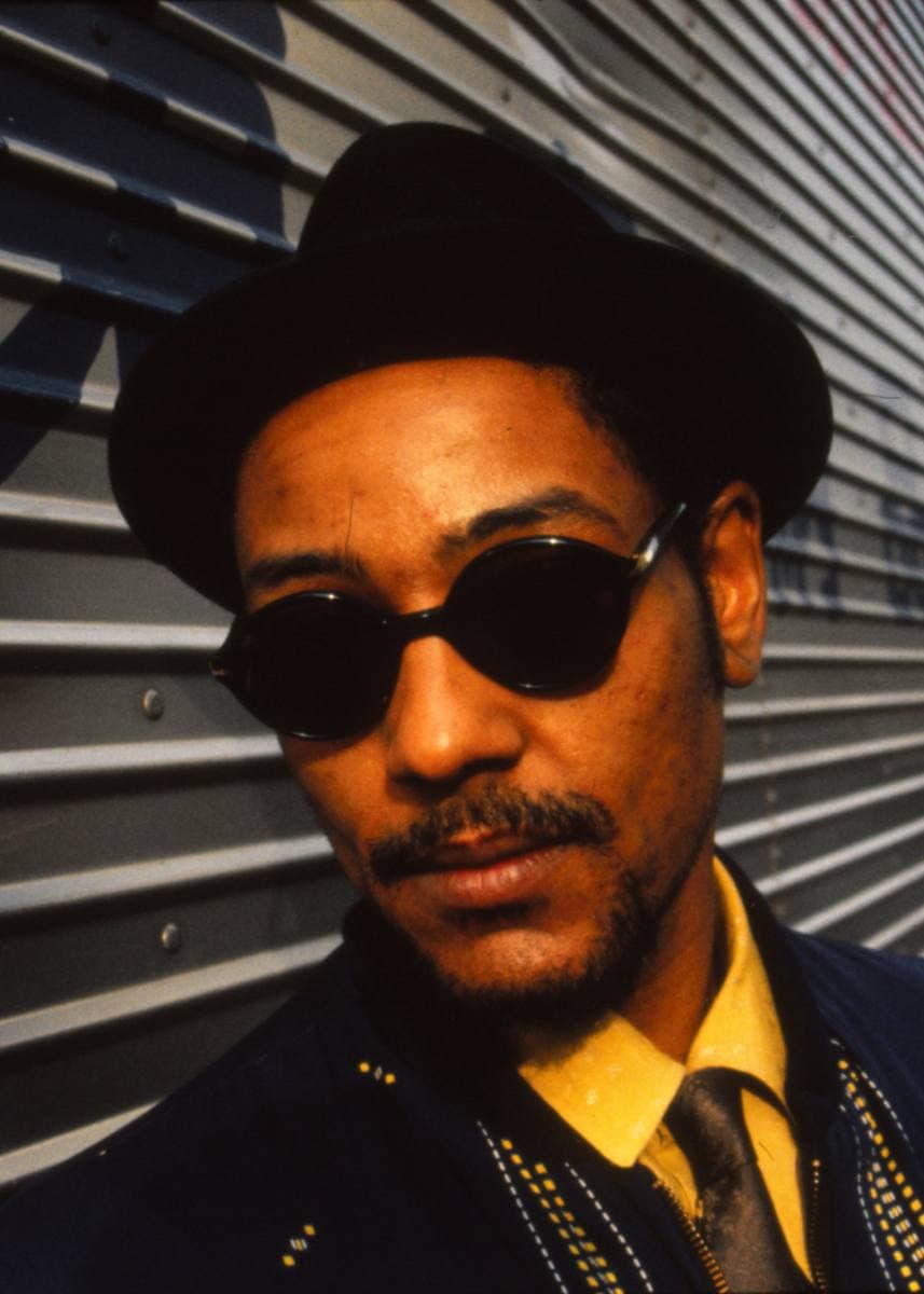 Giancarlo Esposito in a black hat and dark round glasses against a corrugated wall
