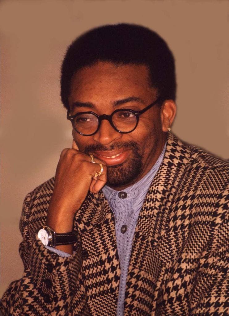 Spike Lee in round glasses and a houndstooth jacket, chin on his hand, smiling
