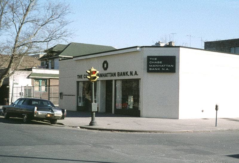 A low white bank building on a Brooklyn street corner, with a 1970s car parked outside