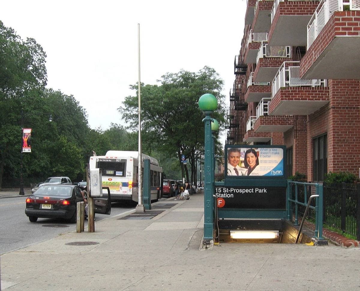 A green subway entrance set into a pavement beside apartment blocks and a bus