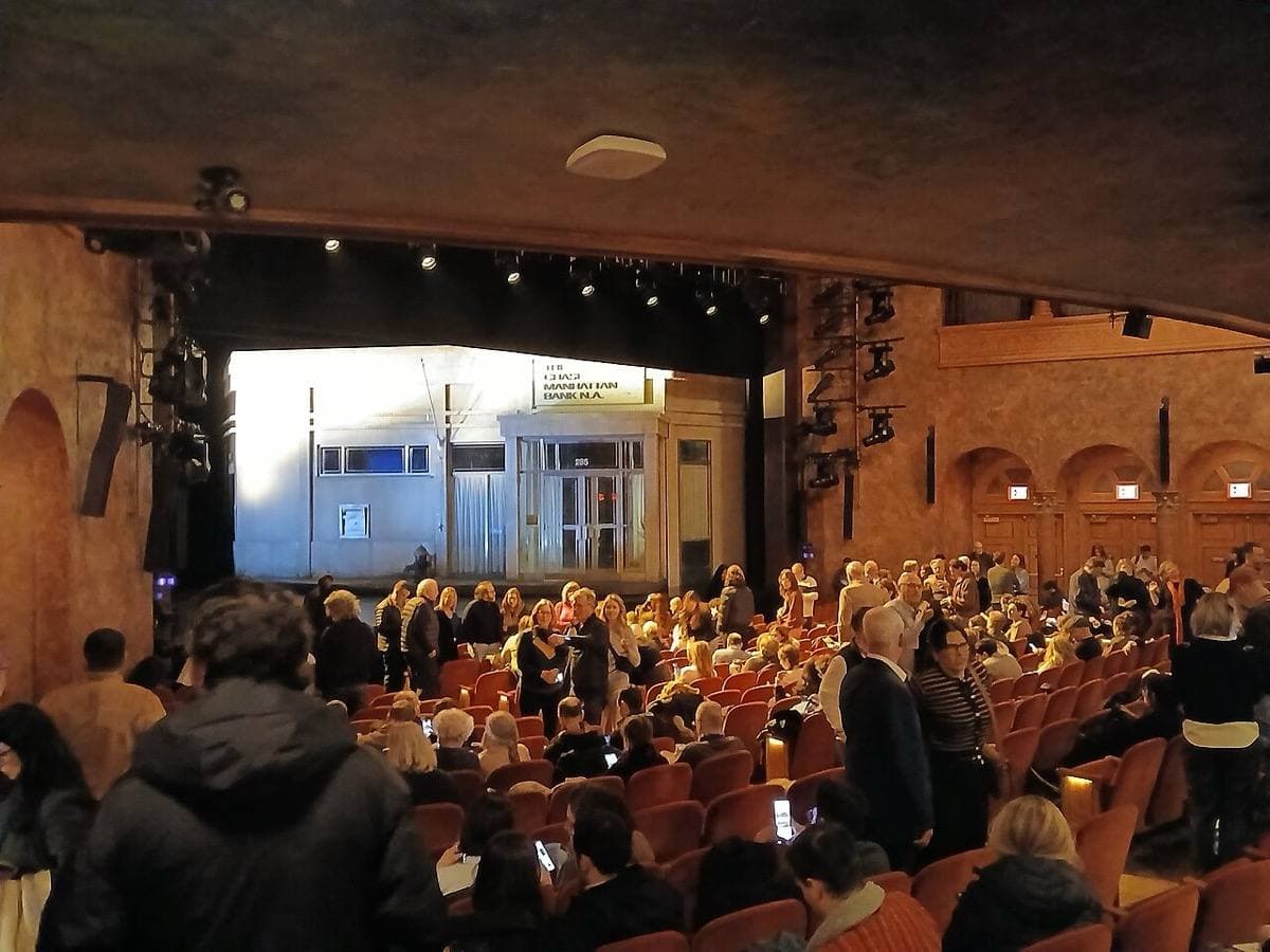 A theatre auditorium full of people, a bank front lit on the stage beyond