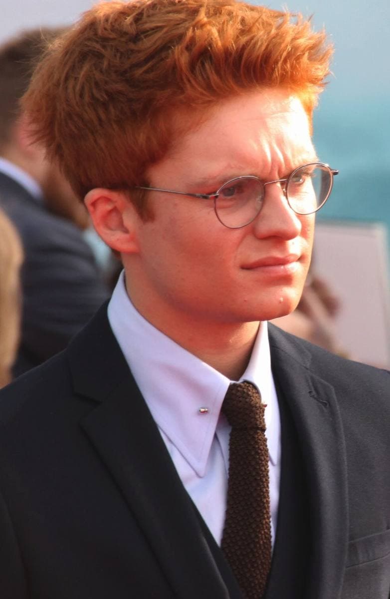 Tom Glynn-Carney, a young man with red hair and round glasses, in a dark suit