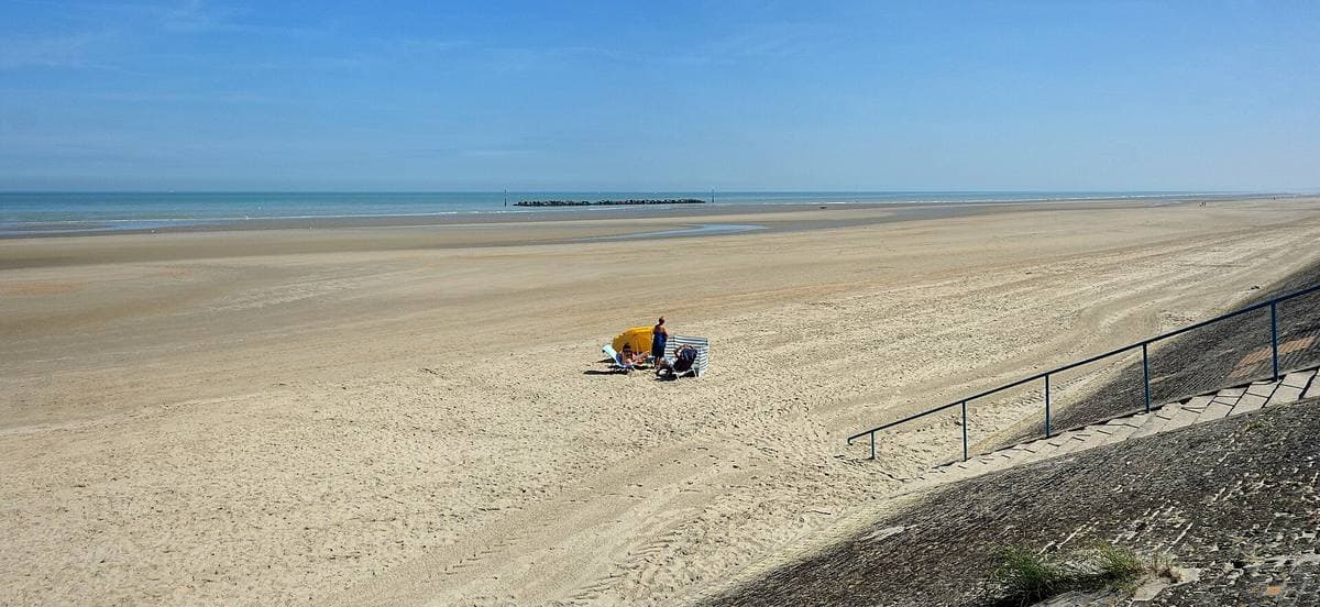 A wide, nearly empty sandy beach under a blue sky, a sea wall and railing on the right