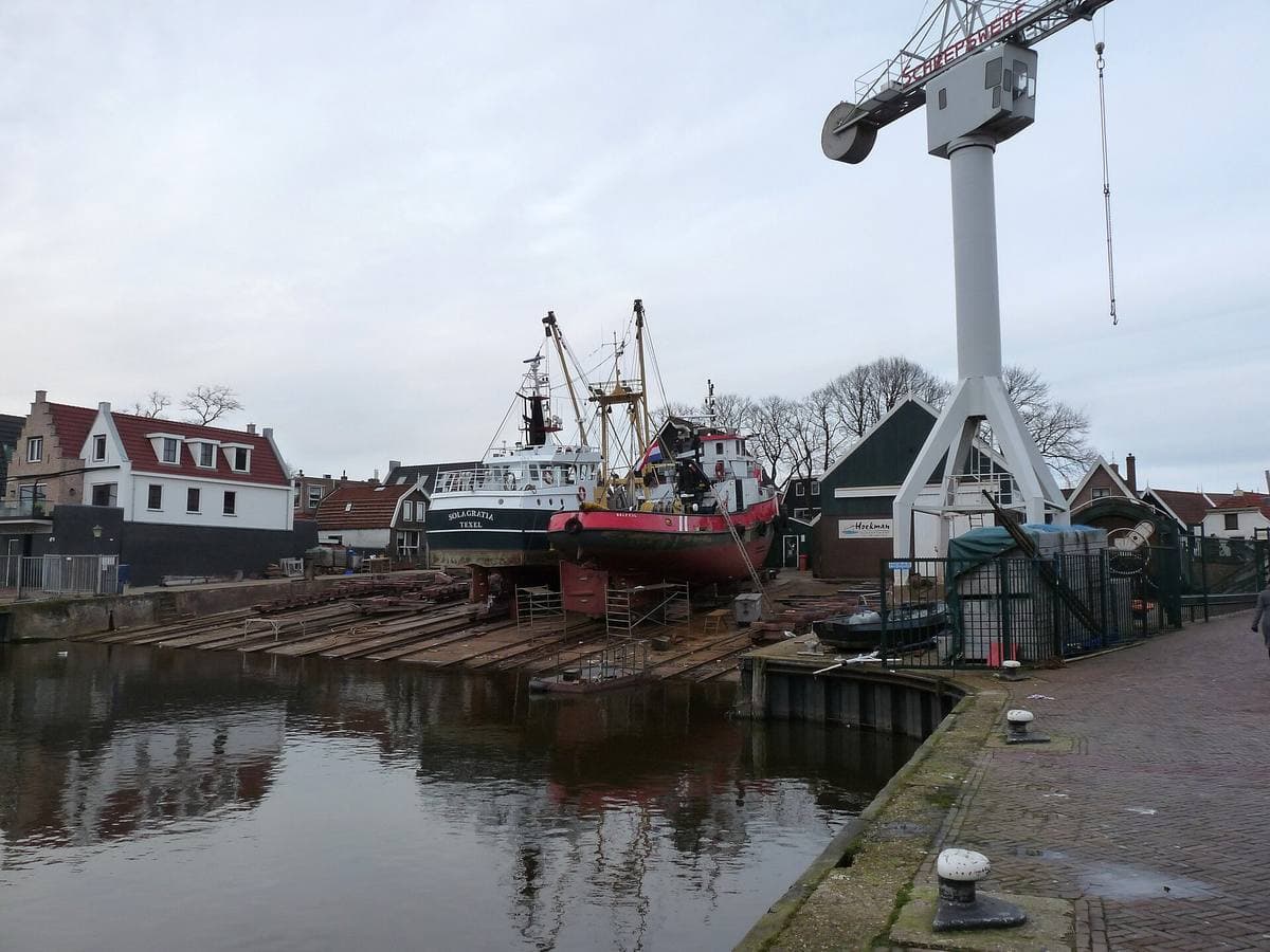A fishing boat on a slipway beside a crane, brick houses across the harbour