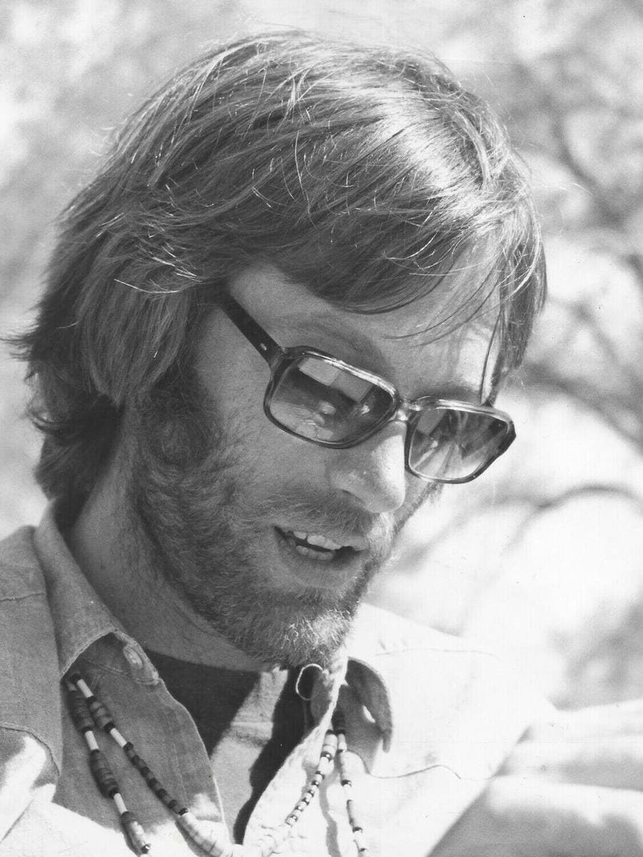 Peter Fonda with long hair, glasses and beads, looking down