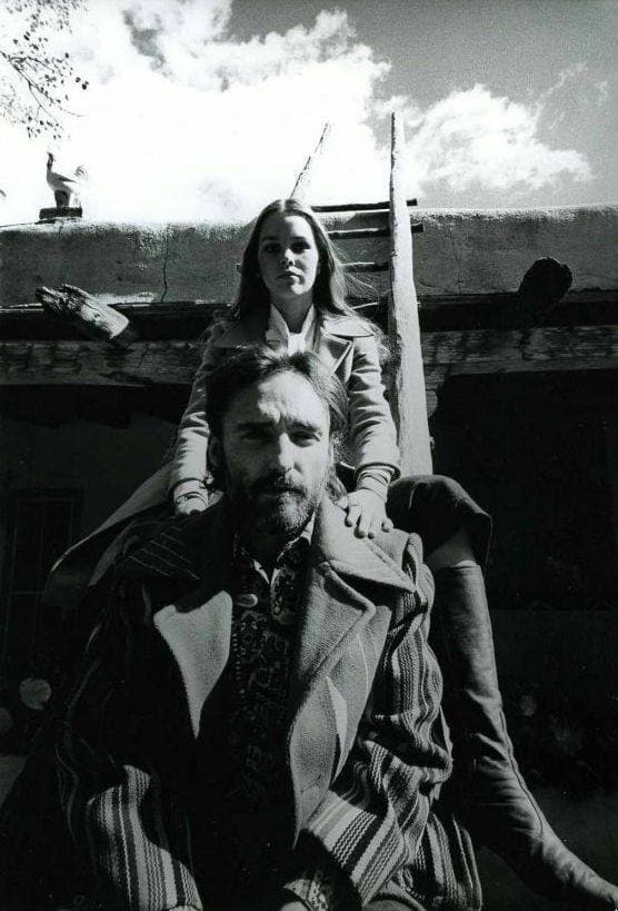 Dennis Hopper seated in a long coat in front of an adobe wall, Michelle Phillips behind him