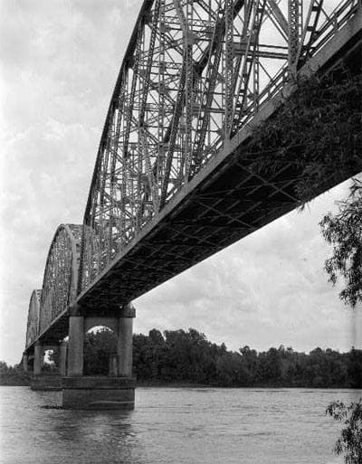 Black-and-white view under a steel truss bridge across a wide river