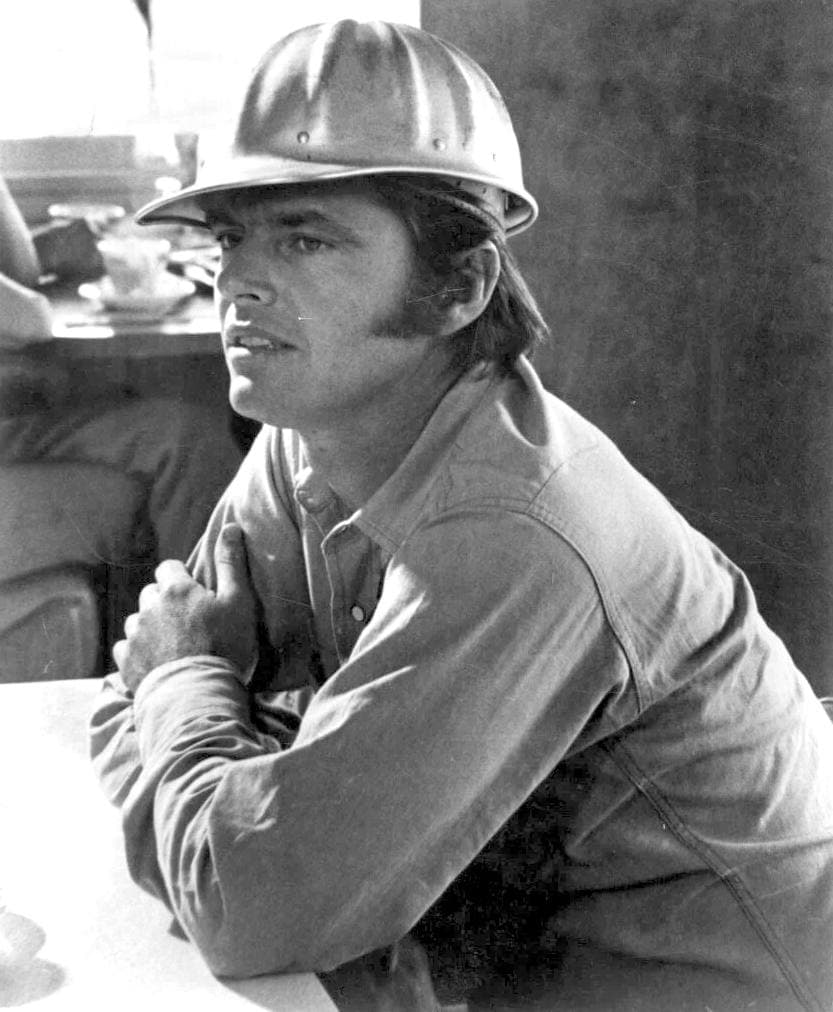 Jack Nicholson in a work shirt and hard hat, leaning on a table