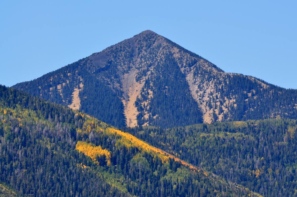 A forested mountain peak with yellow aspen bands under a blue sky