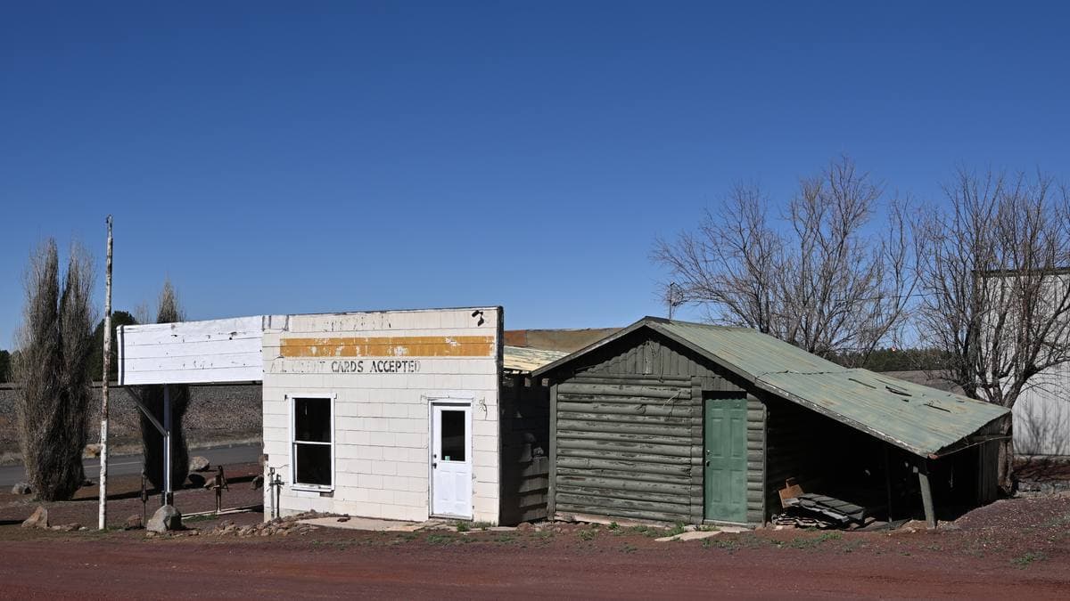 A small white wooden building with a faded painted sign beside a green cabin under a blue sky