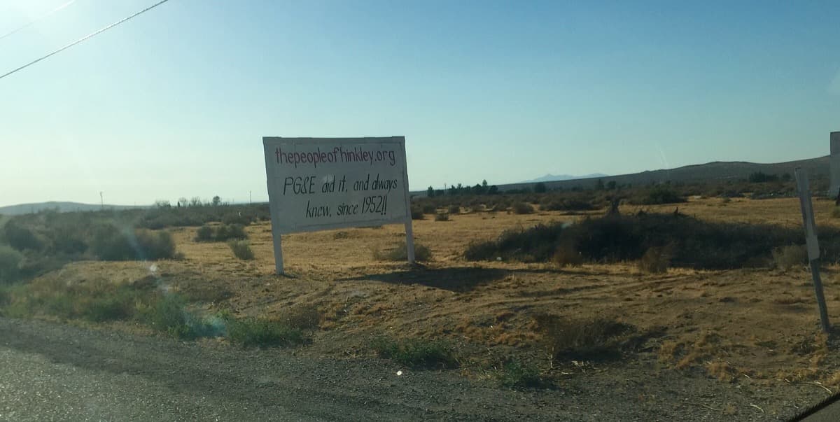 A hand-painted billboard beside a desert road with large lettering accusing a utility company