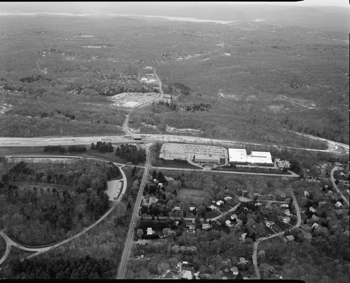 Black-and-white aerial view of woods, fields and a lake beside a parkway