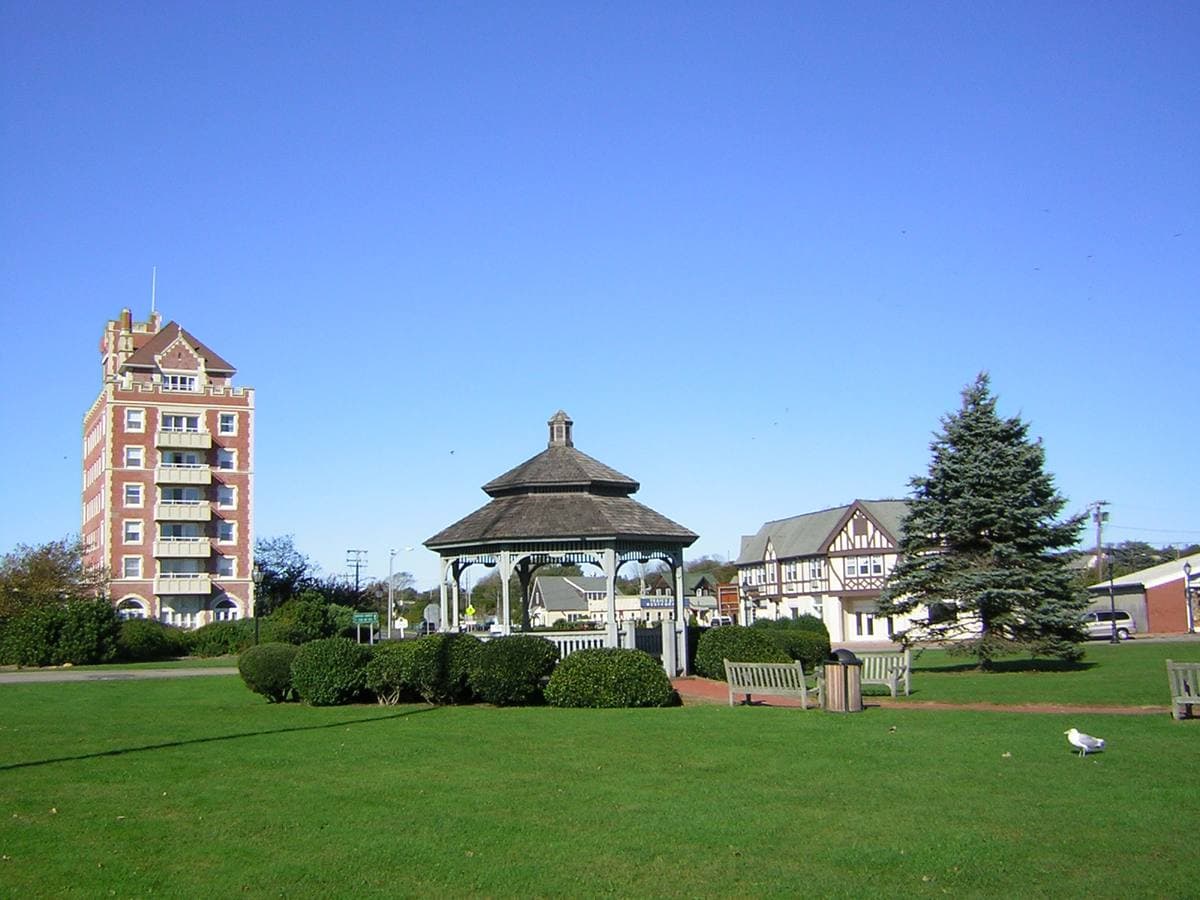 A white gazebo on a green lawn with a tall brick tower behind it