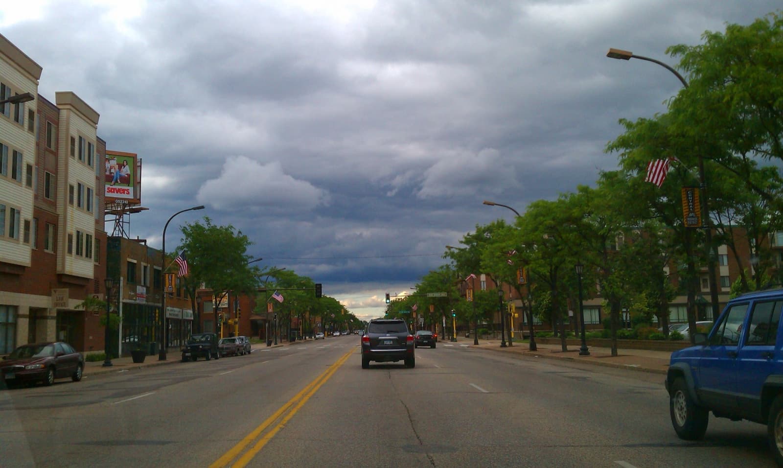 A wide avenue lined with young trees and low buildings under storm clouds