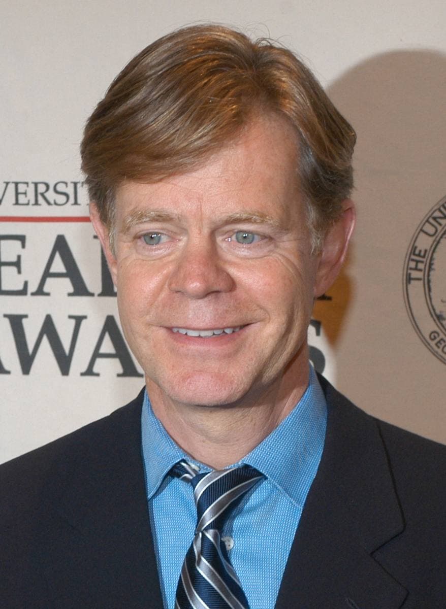 William H. Macy smiling in a dark suit, blue shirt and striped tie