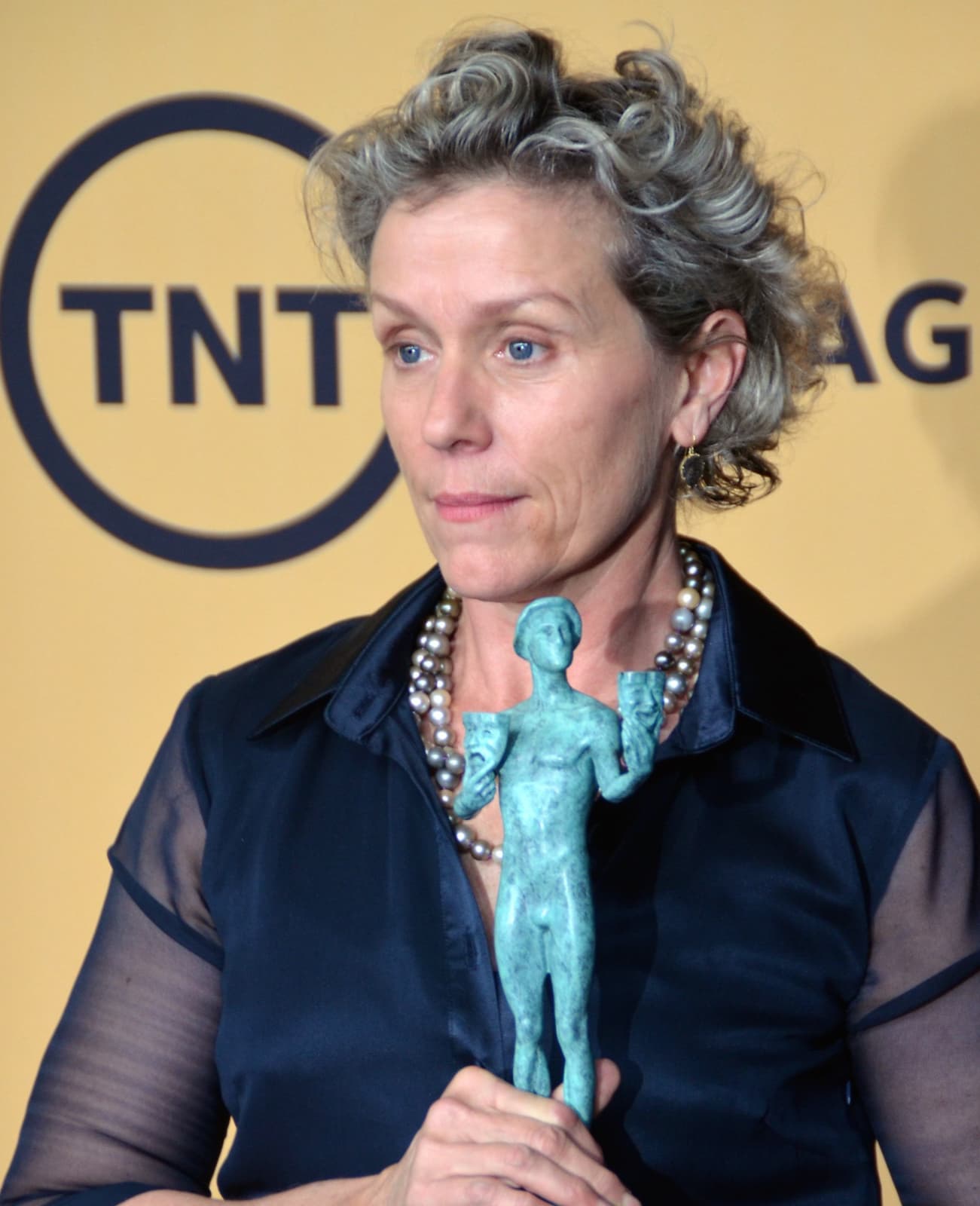 Frances McDormand with curly grey hair, holding a small green statuette