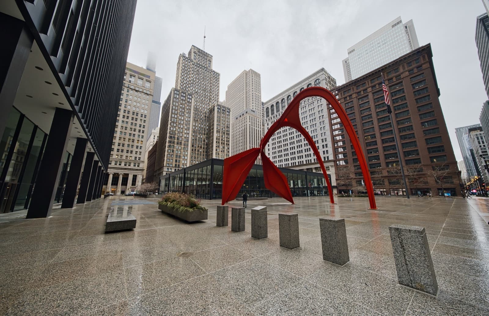 A tall red steel arch sculpture in a granite plaza between black steel towers