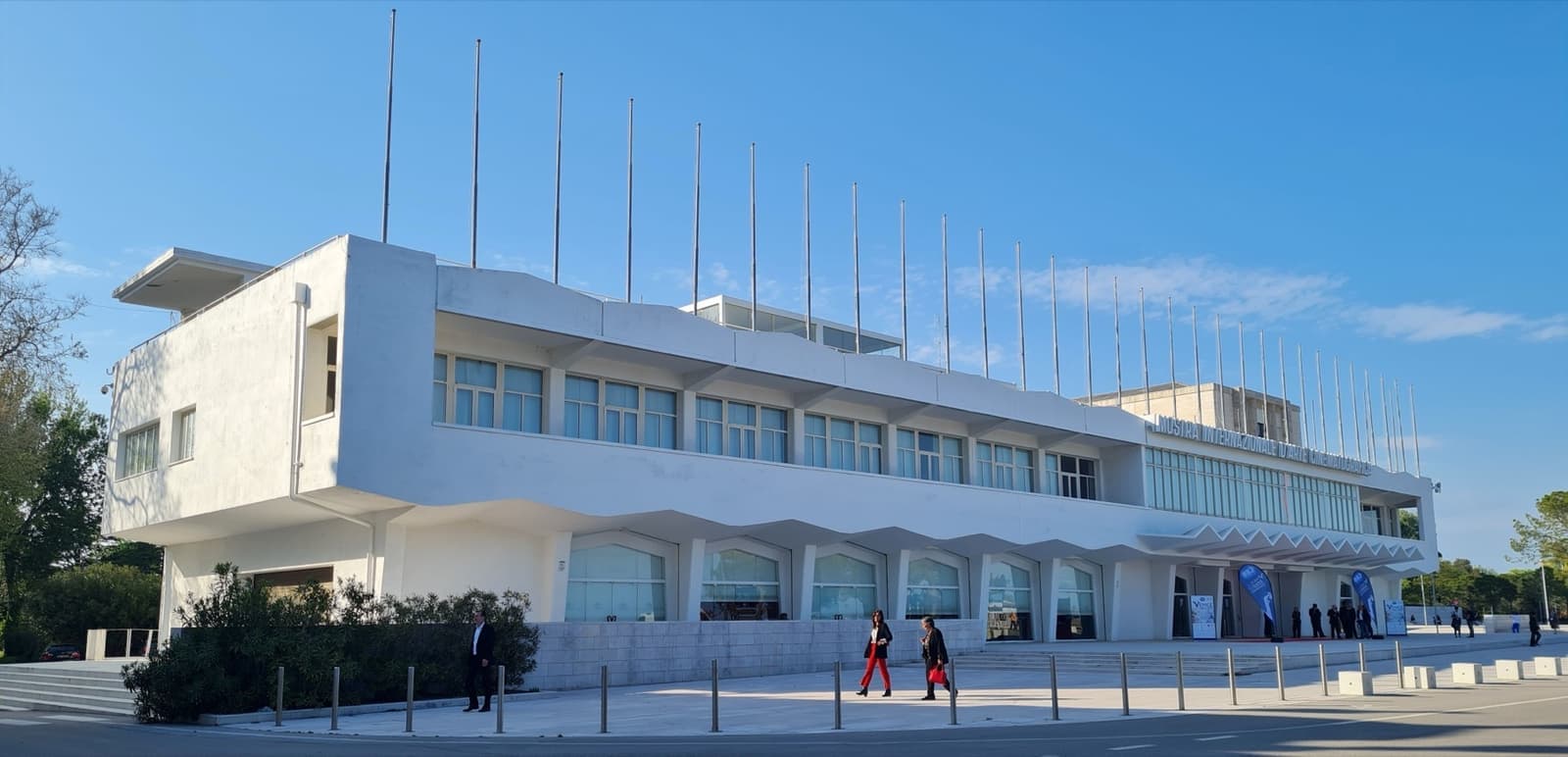 A long white modernist building with flagpoles on its roof, on a wide paved front