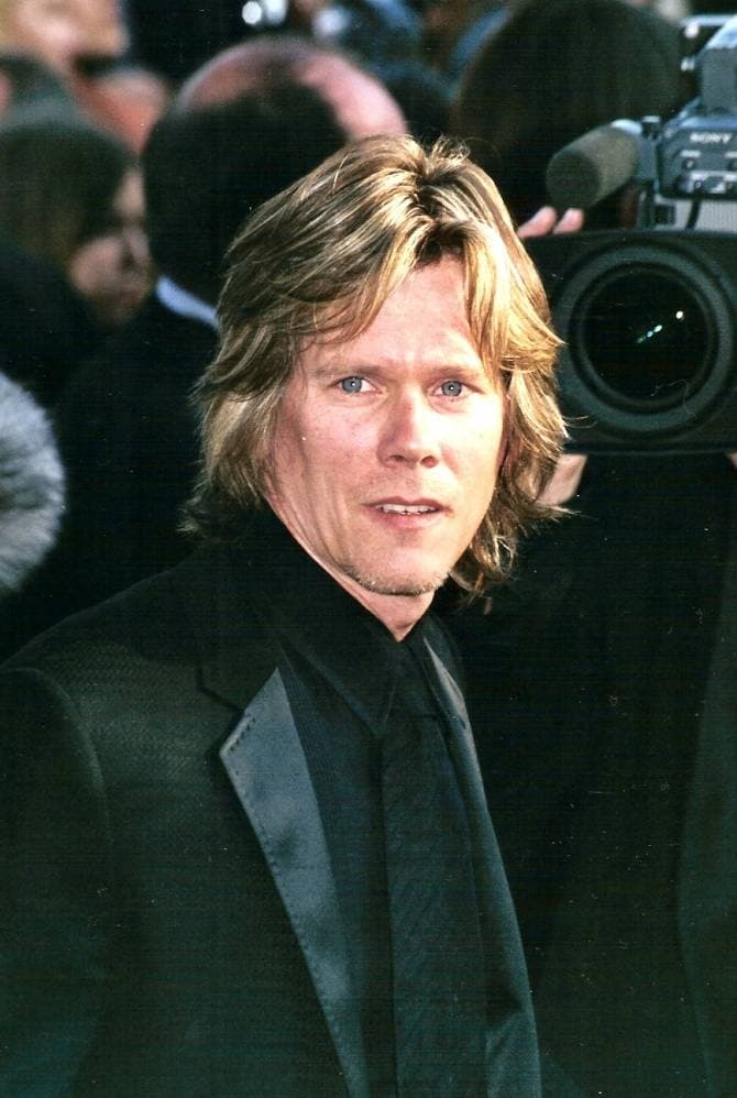 Kevin Bacon with shoulder-length fair hair in a dark suit, photographers behind him