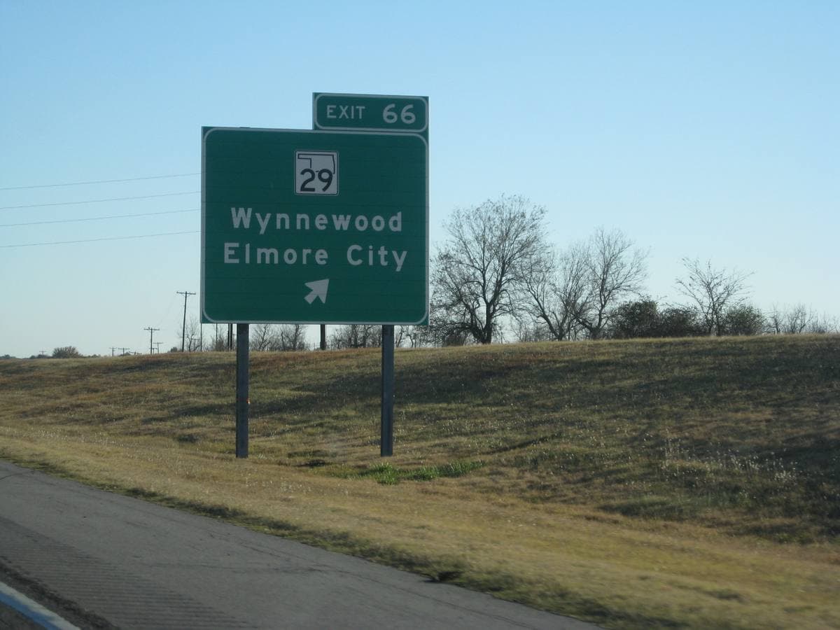 A green motorway exit sign for Wynnewood and Elmore City on a dry grass verge