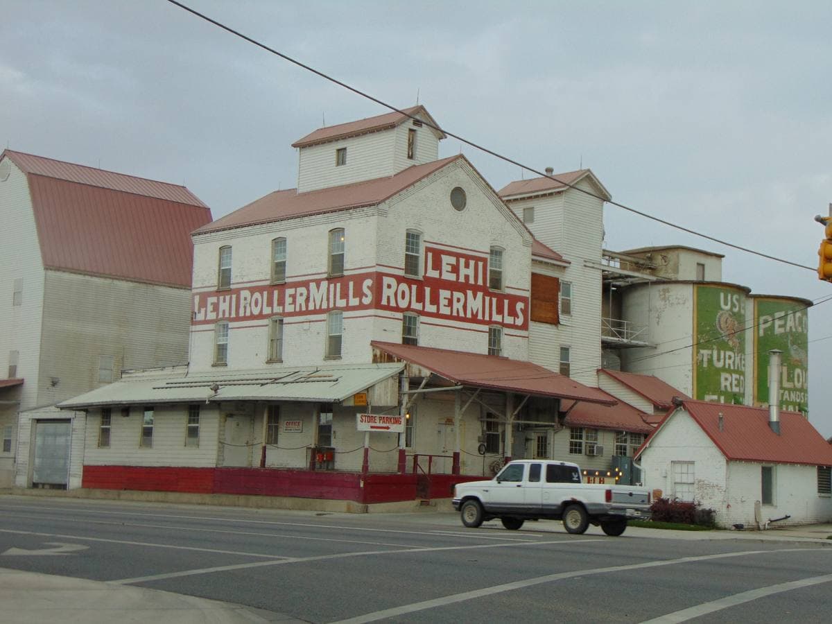 A tall white wooden mill with red trim and painted lettering, silos behind, a street in front