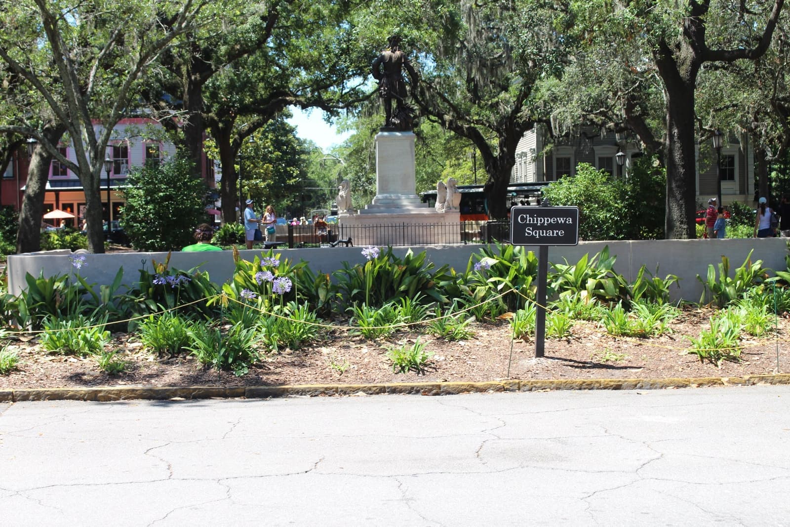 A park square under live oaks with a bronze statue on a plinth and a small Chippewa Square sign in a flower bed