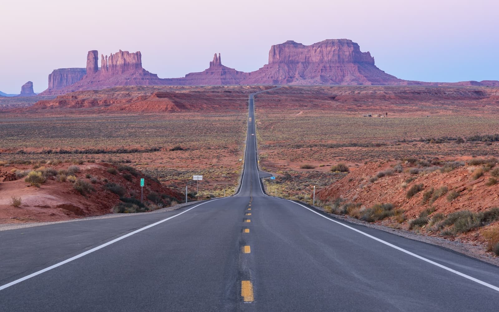 A straight two-lane road running towards red buttes under a pink dawn sky