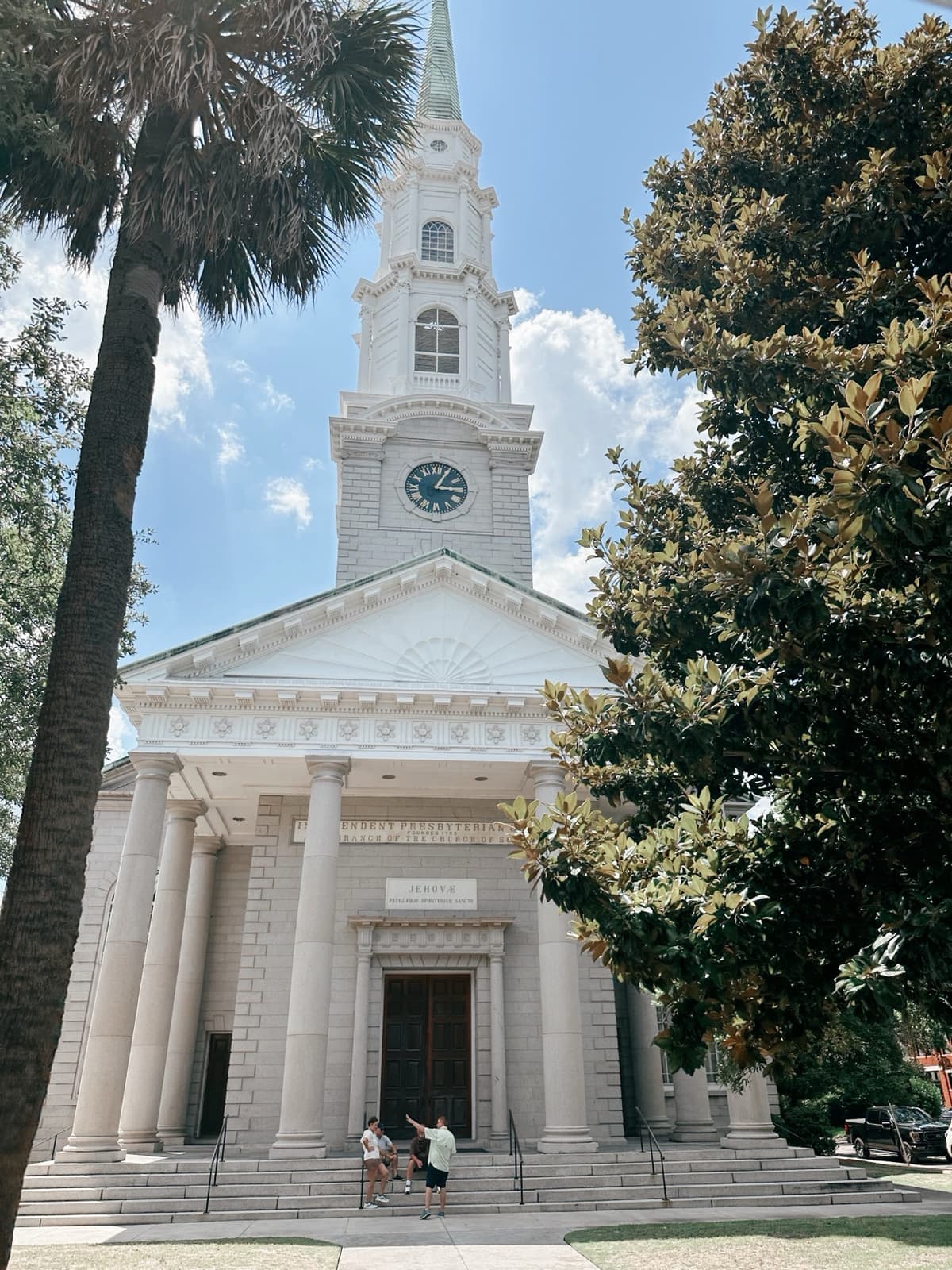 A white church with columns and a tall steeple, framed by a palm tree