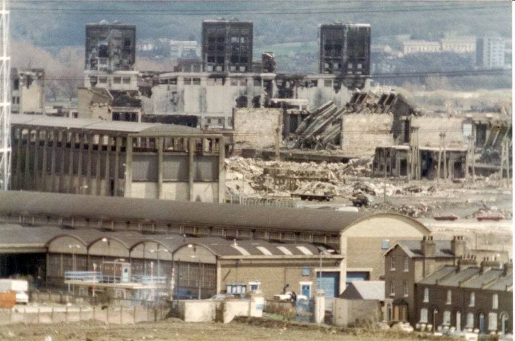 Wide view over roofless brick and concrete gasworks buildings, with tall blocks behind