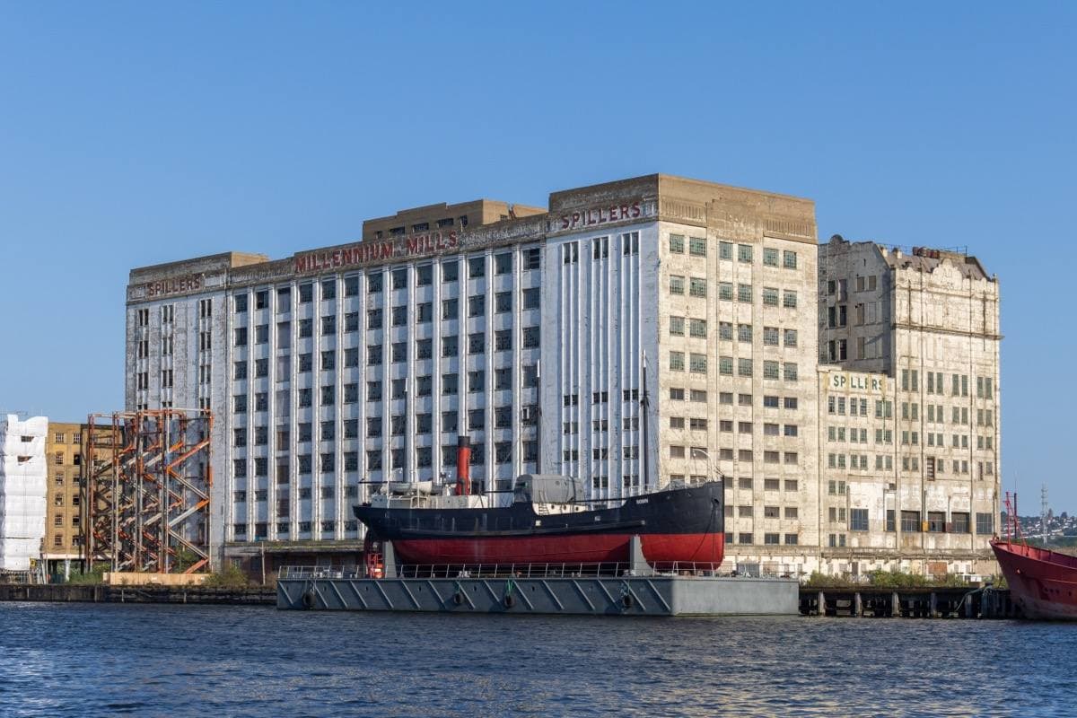 A tall pale concrete mill building across the dock, a small steamship moored before it