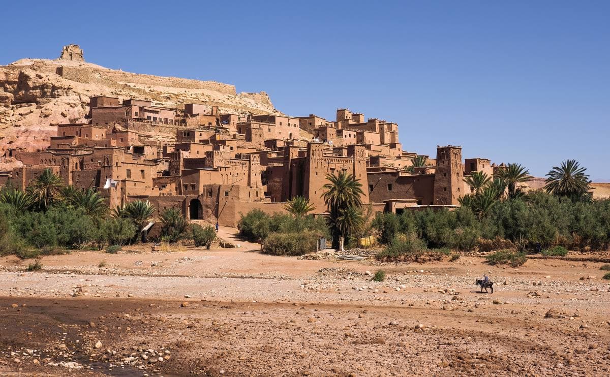 Red earth towers and houses climbing a hill above palms and a dry riverbed
