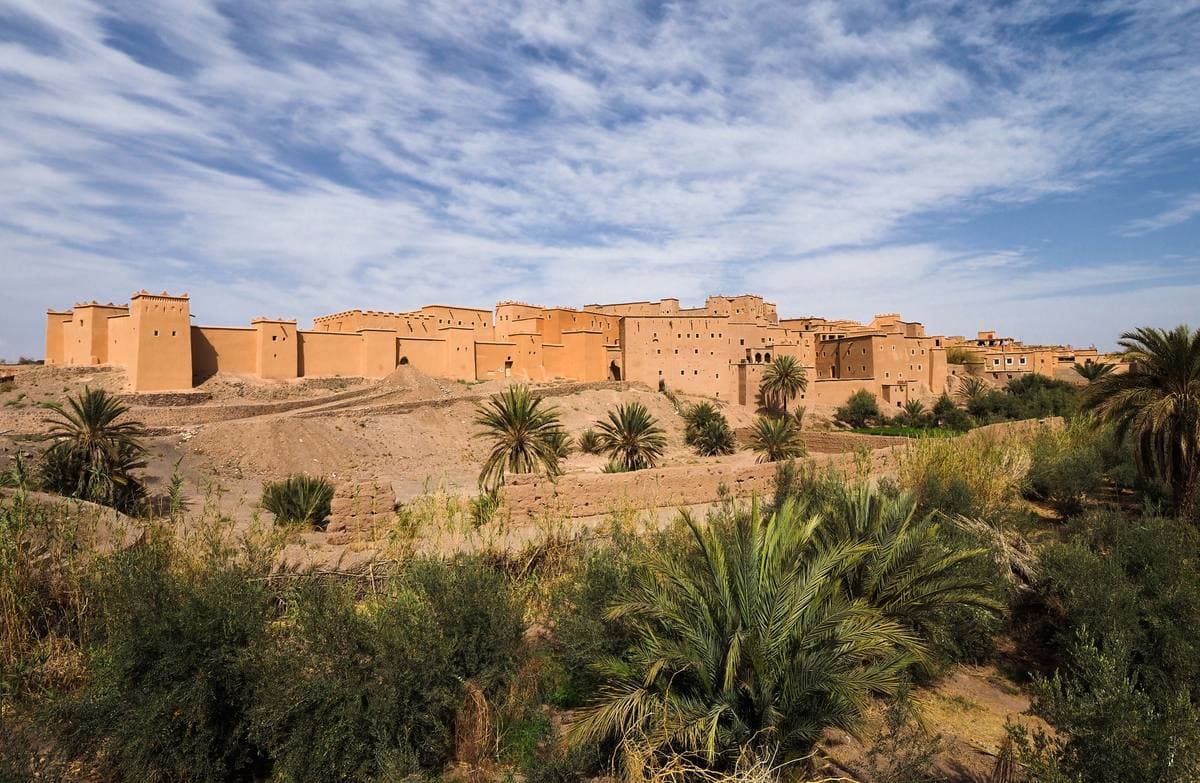 A long earth-red kasbah behind palm trees under a blue sky