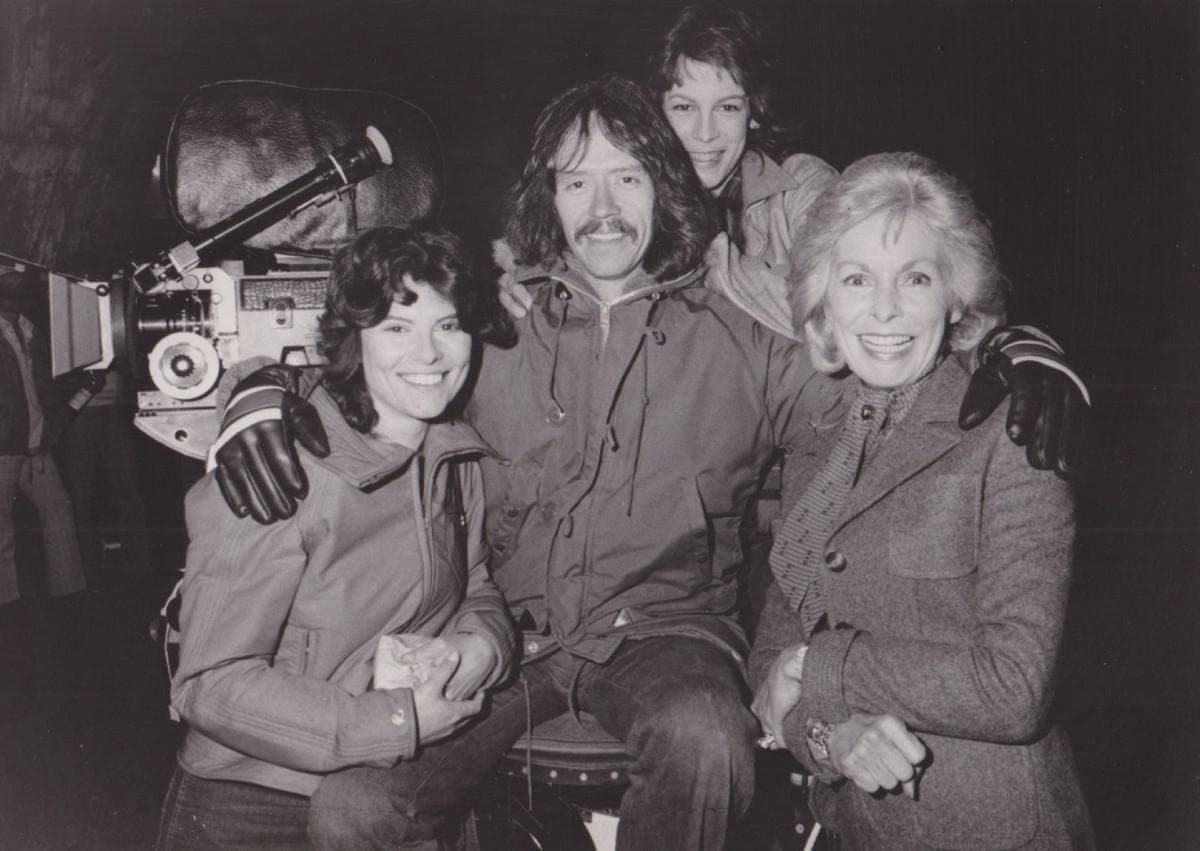A black-and-white press photograph of a bearded John Carpenter beside a film camera with three smiling women
