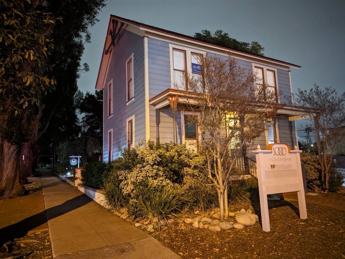 A pale blue two-storey clapboard house with a porch, lit at dusk behind young trees, with a small sign on the lawn