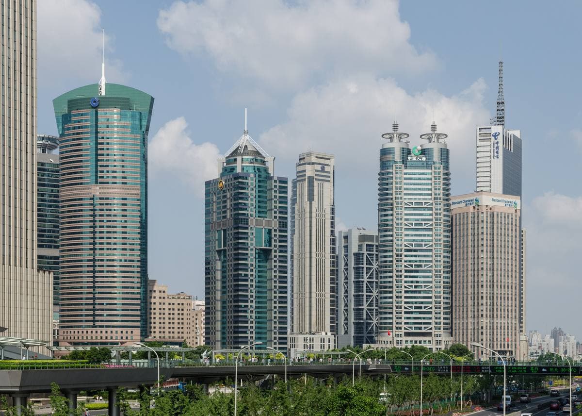 Green-glass skyscrapers in Lujiazui seen from a raised walkway, trees and a road below