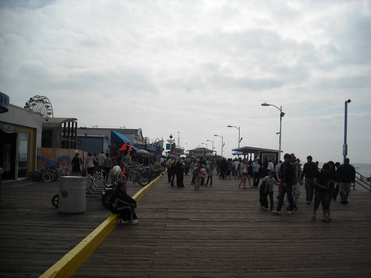 People walking on the wooden deck of Santa Monica Pier under a grey sky, a Ferris wheel behind