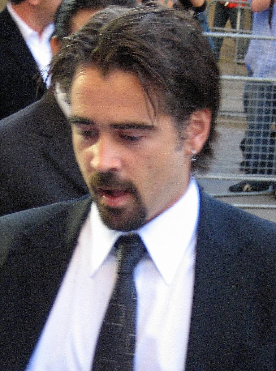 Colin Farrell, dark hair swept back, in a dark suit and tie, looking down