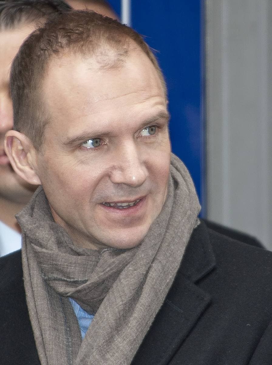 Ralph Fiennes in a grey scarf and dark coat, smiling slightly