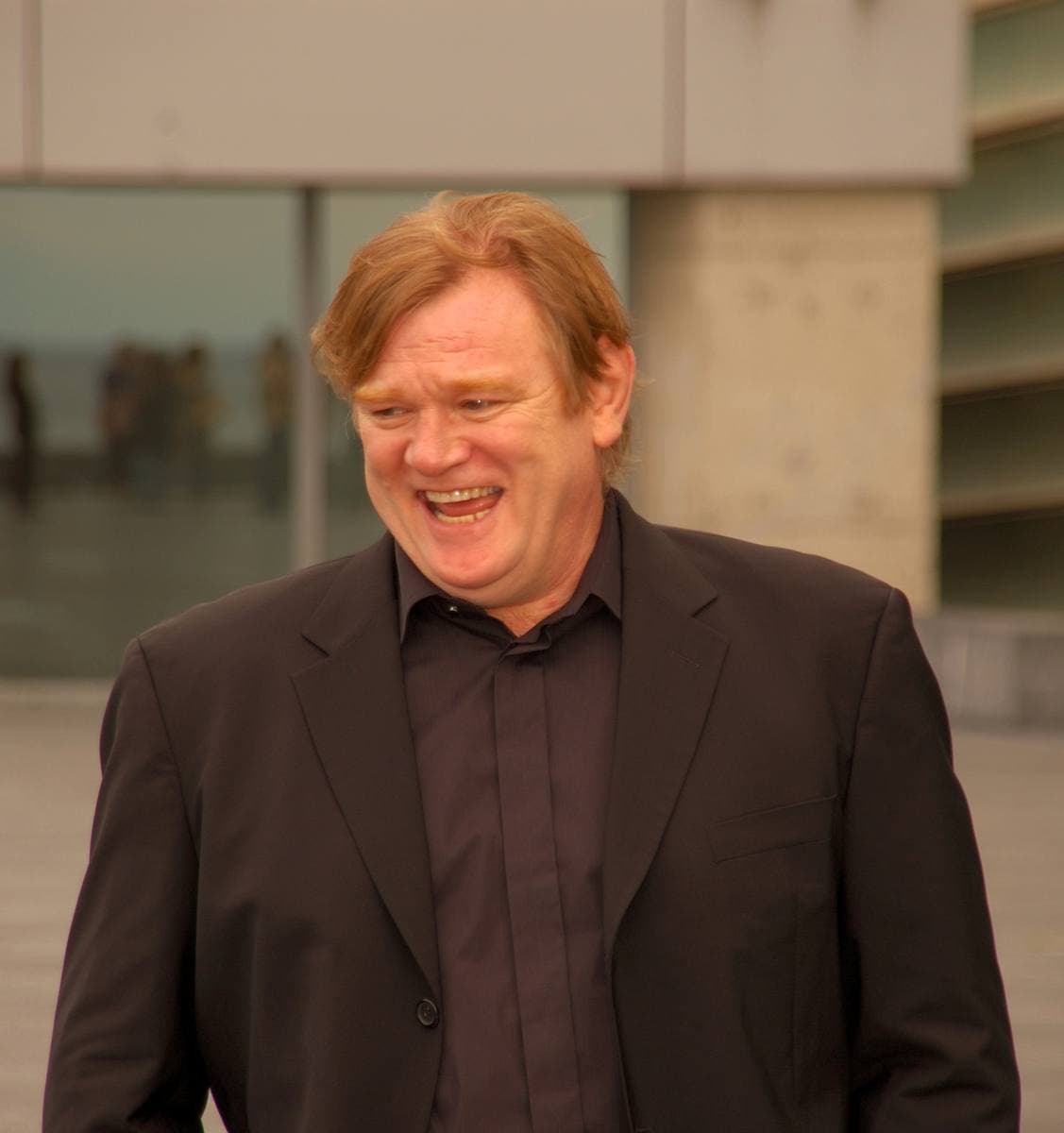 Brendan Gleeson, laughing, in a dark jacket and open-necked shirt
