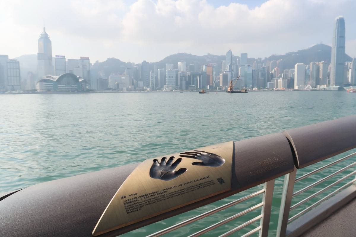A bronze handprint plaque on a curved harbour rail, the Hong Kong Island skyline beyond
