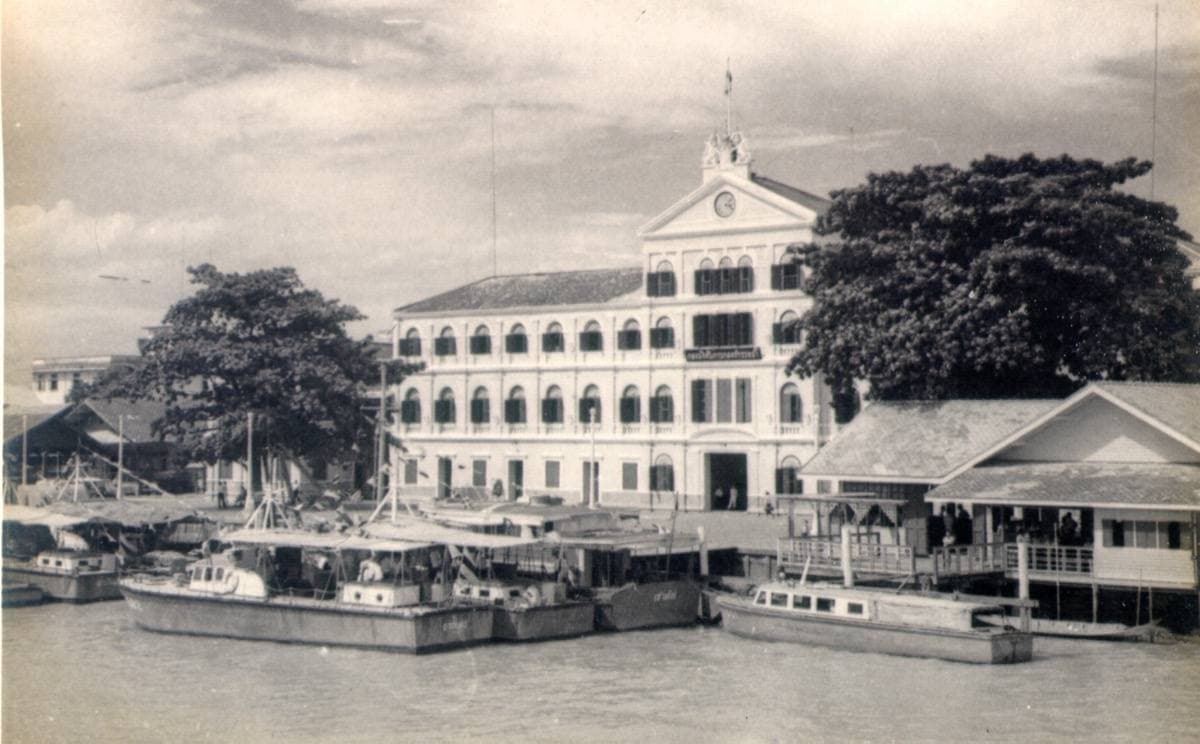 A black-and-white view of a three-storey colonial building above boats on a river