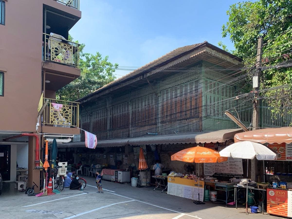 A long two-storey wooden building on a narrow lane, umbrellas and a food stall in front