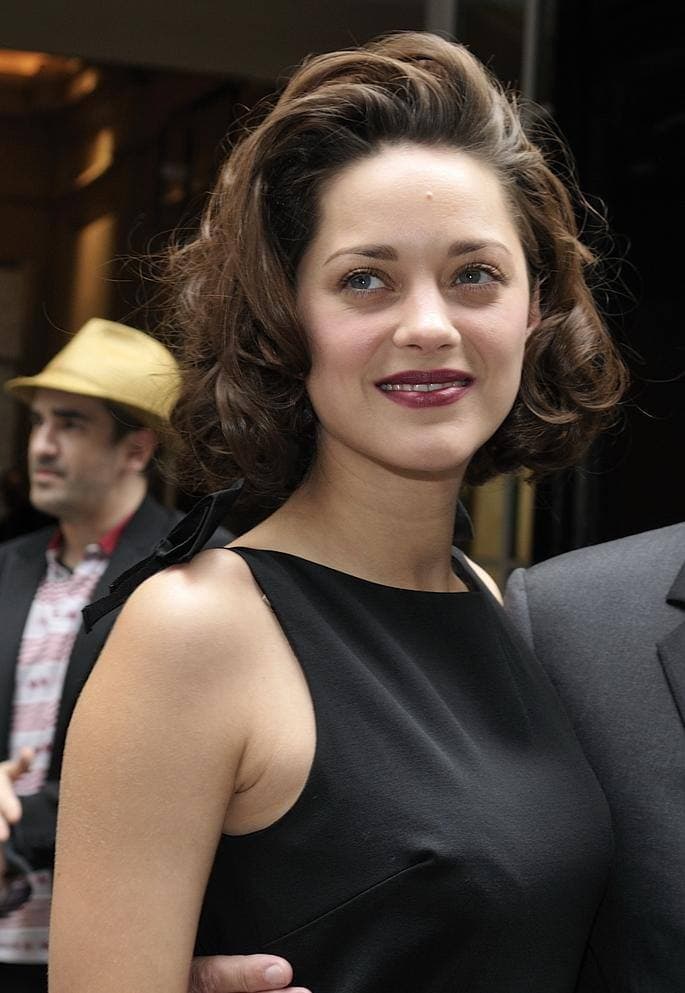 Marion Cotillard with short curled hair in a black sleeveless dress, smiling