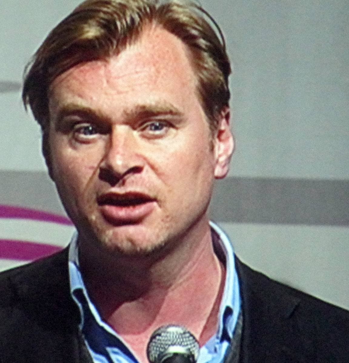 Christopher Nolan in a dark jacket and blue shirt speaking into a microphone