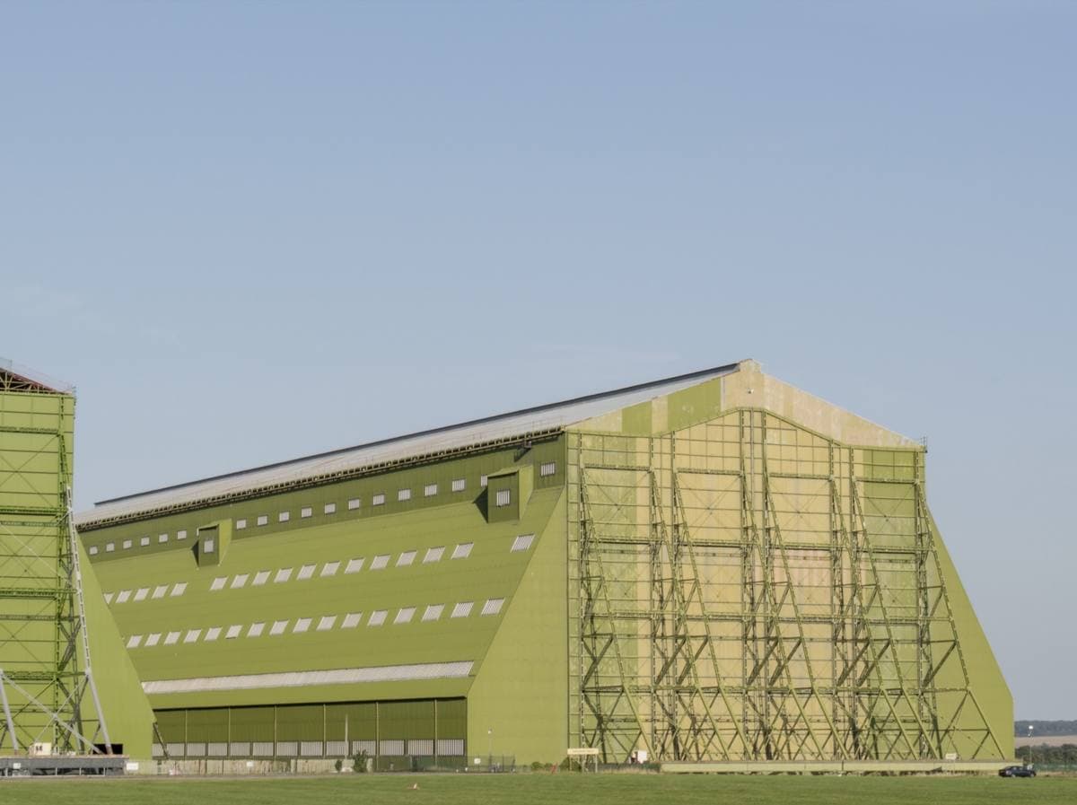 A huge green airship hangar with scaffolding along one side, standing in a field