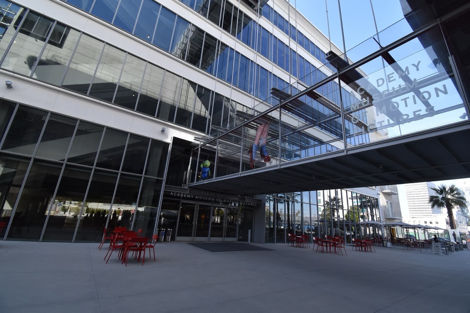 A glass-fronted museum building with a footbridge overhead and red chairs outside