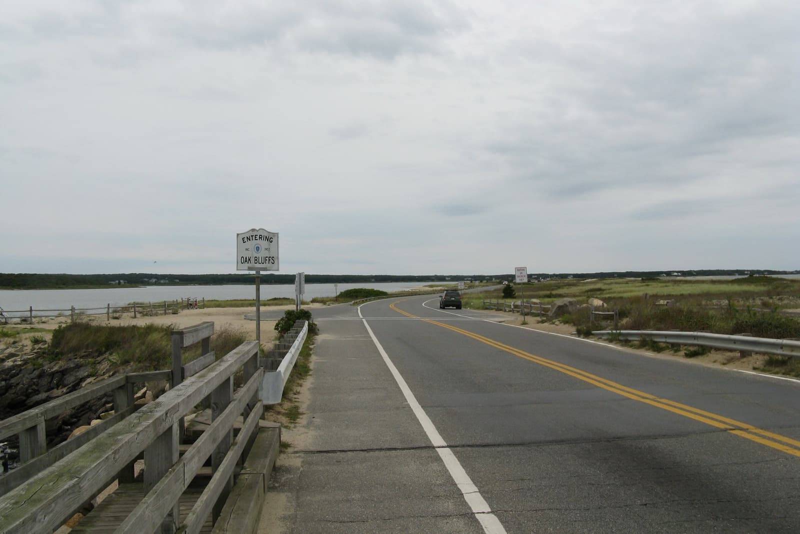 A two-lane road on a causeway with a wooden bridge rail and a town sign